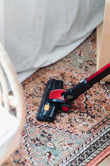 A close-up image of a handheld vacuum cleaner with a black and red design, positioned on a traditional patterned rug with intricate designs in shades of red, beige, and blue. The vacuum appears to be actively cleaning a carpeted surface in a domestic setting, with part of a white curtain or fabric background and light wooden furniture visible nearby. The surface is clean and vacuumed, highlighting the importance of surface cleaning and deep cleaning for maintaining hygiene in Wimbledon Village homes. Merton Carpet Cleaning provides professional domestic cleaning services to ensure thorough sanitisation and upkeep of carpets and rugs.