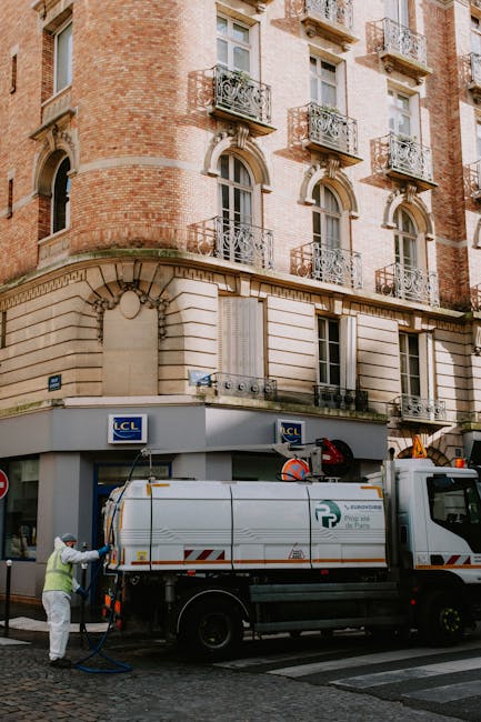 A street-level view of a multi-story building with a classic brick façade and ornate metal balconies on each window, situated on Morden High Street (SM4). The ground floor features large windows with beige stone trims and a corner shop with a grey sign displaying the LCL logo. In front of the building, a white maintenance truck equipped with tools and hoses is parked on the cobbled street. A worker wearing a white protective suit and high-visibility vest is operating equipment connected to the truck, likely conducting cleaning or sanitisation tasks. The scene is illuminated by natural daylight, and the overall setting suggests routine building or street maintenance, emphasizing cleanliness and upkeep consistent with professional cleaning standards. Merton Carpet Cleaning offers expert surface cleaning and deep cleaning services, contributing to hygiene and property maintenance in the local area.