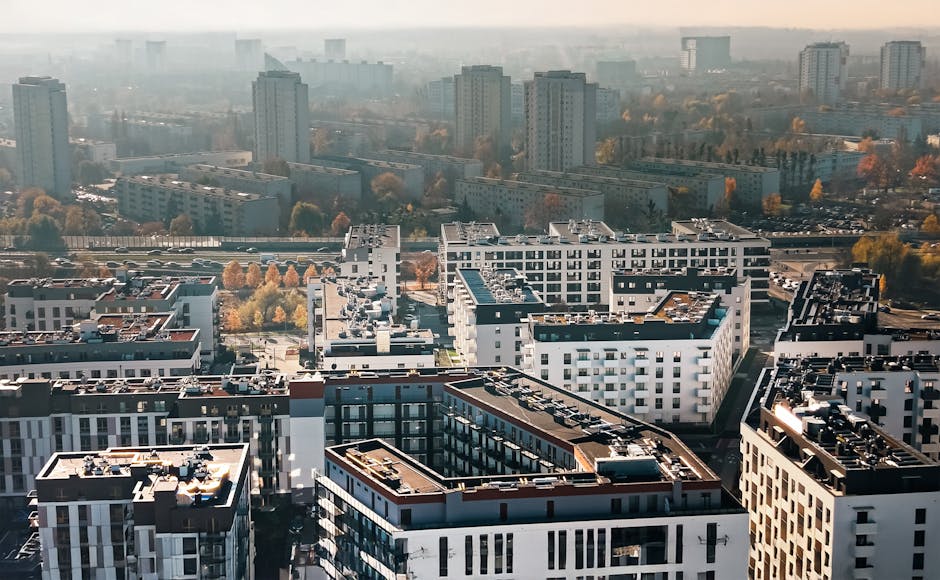Aerial view of a modern residential complex in Colliers Wood Flats, Merton, featuring white and gray multi-story buildings with flat rooftops, some equipped with black and silver HVAC units, and large windows reflecting sunlight. The foreground shows the rooftops with visible vents, mechanical systems, and walkways, while the background reveals tall, distant apartment blocks and tree-lined streets with autumn foliage. The scene is well-lit with natural daylight, highlighting the clean, maintained surfaces of the buildings. Merton Carpet Cleaning emphasizes surface cleaning and sanitisation in maintaining the hygienic condition of such residential properties.
