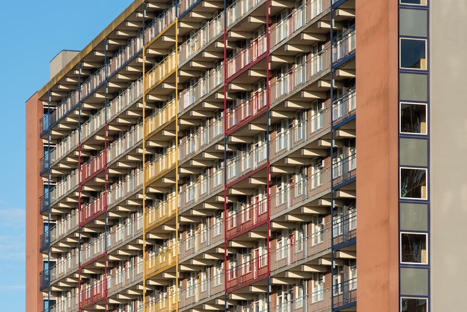 Close-up view of a multi-storey residential building's exterior showcasing numerous apartment balconies arranged in a grid pattern. Each balcony features metal railings painted in alternating colors of red, yellow, and blue, with transparent glass panels and white support structures. The building facade displays modern architectural design with a mix of beige and orange-toned walls, and large rectangular windows reflecting the sky. The image highlights clean, well-maintained surfaces with no visible dirt or clutter, emphasizing the importance of surface cleaning and maintenance in residential environments. Visible is a section of the building's flat roof and the overall structure benefits from natural daylight, accentuating the neat appearance of the balconies and external walls. This visual aligns with the theme of sanitation and surface cleaning relevant to estate cleaning for flats, as discussed in the Estate Cleaning Guide for Colliers Wood Flats, MERTON by Merton Carpet Cleaning.