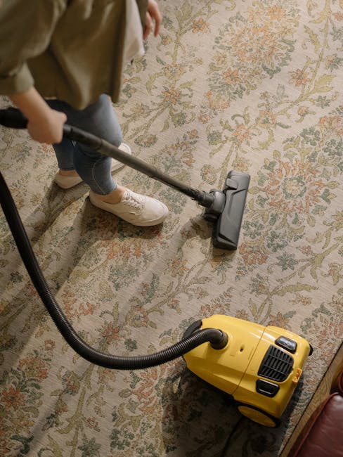 Close-up of a person using a yellow and black vacuum cleaner for surface cleaning on a patterned carpet. The person's hands are holding a cloth to clean the interior of the vacuum's dust container, wh