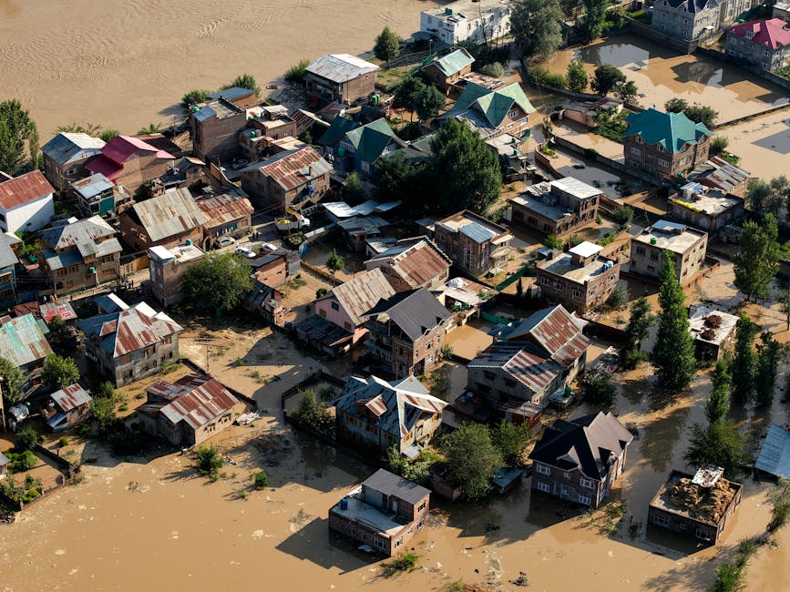 A flooded residential area in Merton during heavy rain, with muddy water covering the front garden and driveway of a two-story house featuring a tiled roof and white walls. The property is surrounded